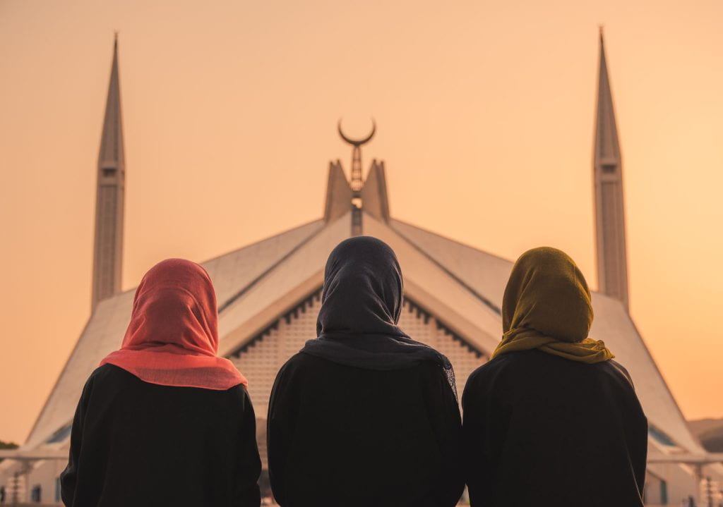 Three women wearing colored head scarves looking at a mosque in the distance with their backs towards the viewer. 